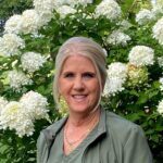A smiling woman with blonde hair stands in front of white hydrangea flowers.